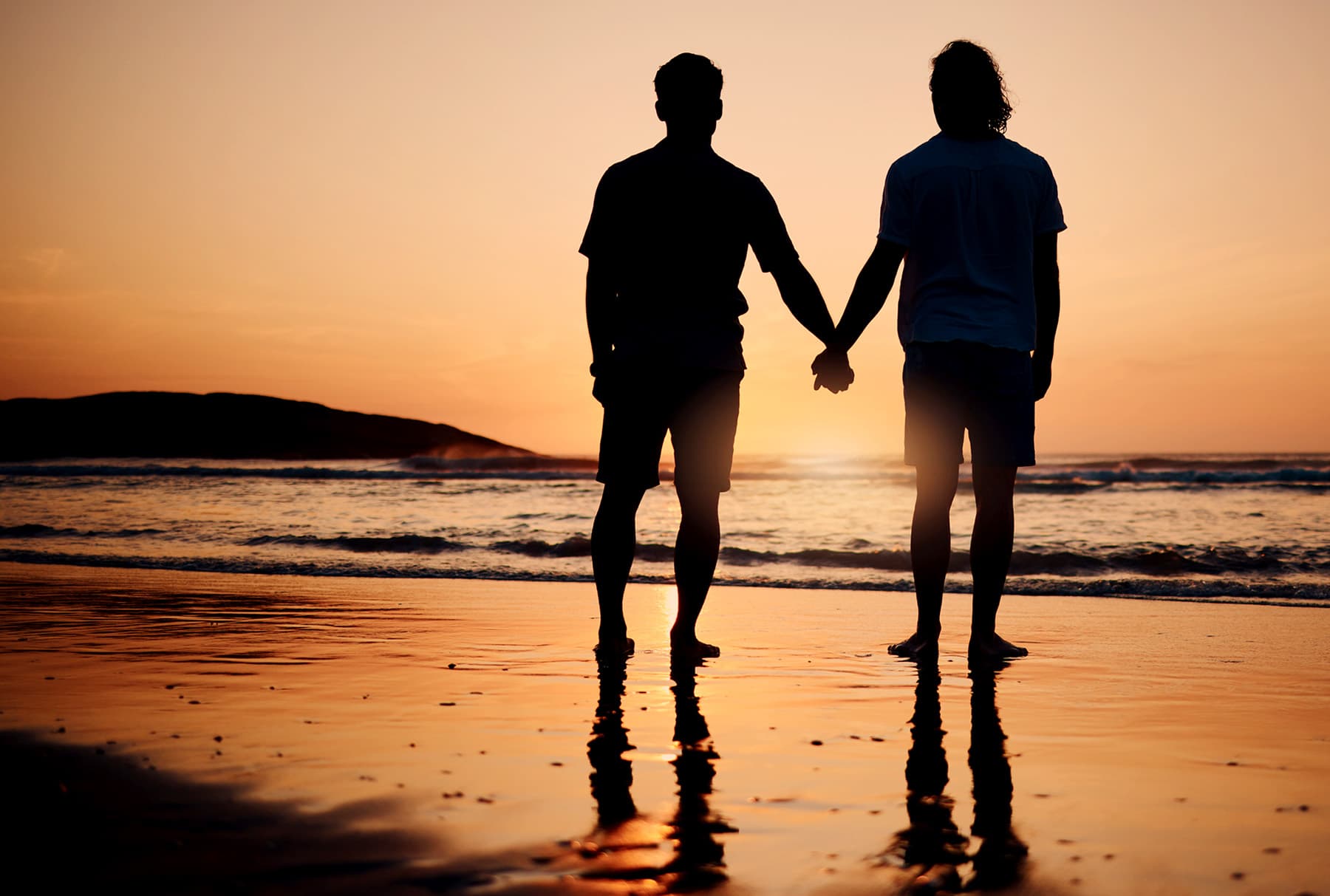 Two people standing hand in hand at the water's edge at sunset, seen from behind in silhouette, their reflections on the wet sand