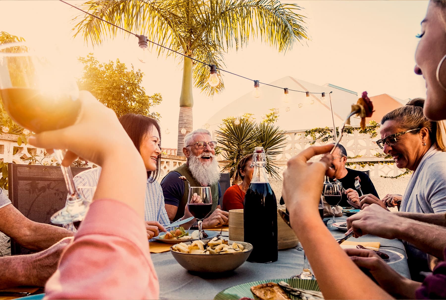 A long table of people at an outdoor dinner under string lights at sunset, glasses raised mid-conversation