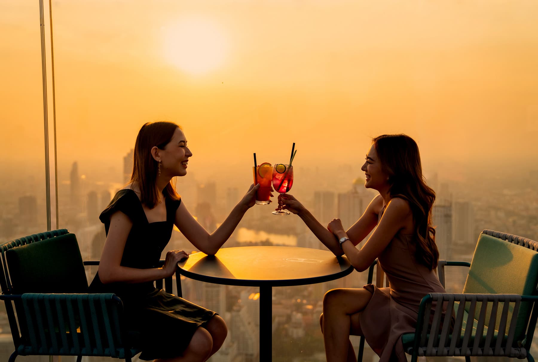 Two people at a rooftop table raising drinks to each other, a hazy city skyline far below at sunset