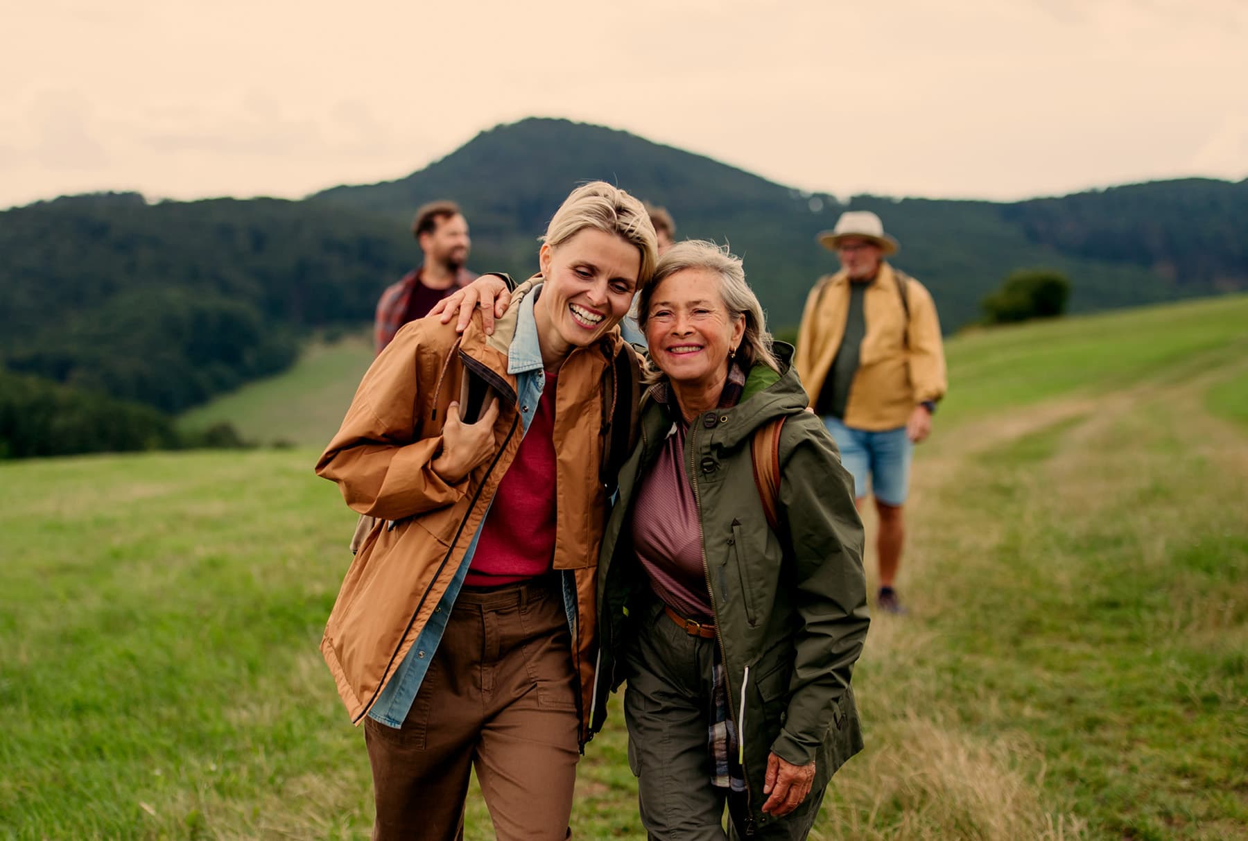 Two women walking arm in arm and laughing on a grassy hillside, two more of their group following behind