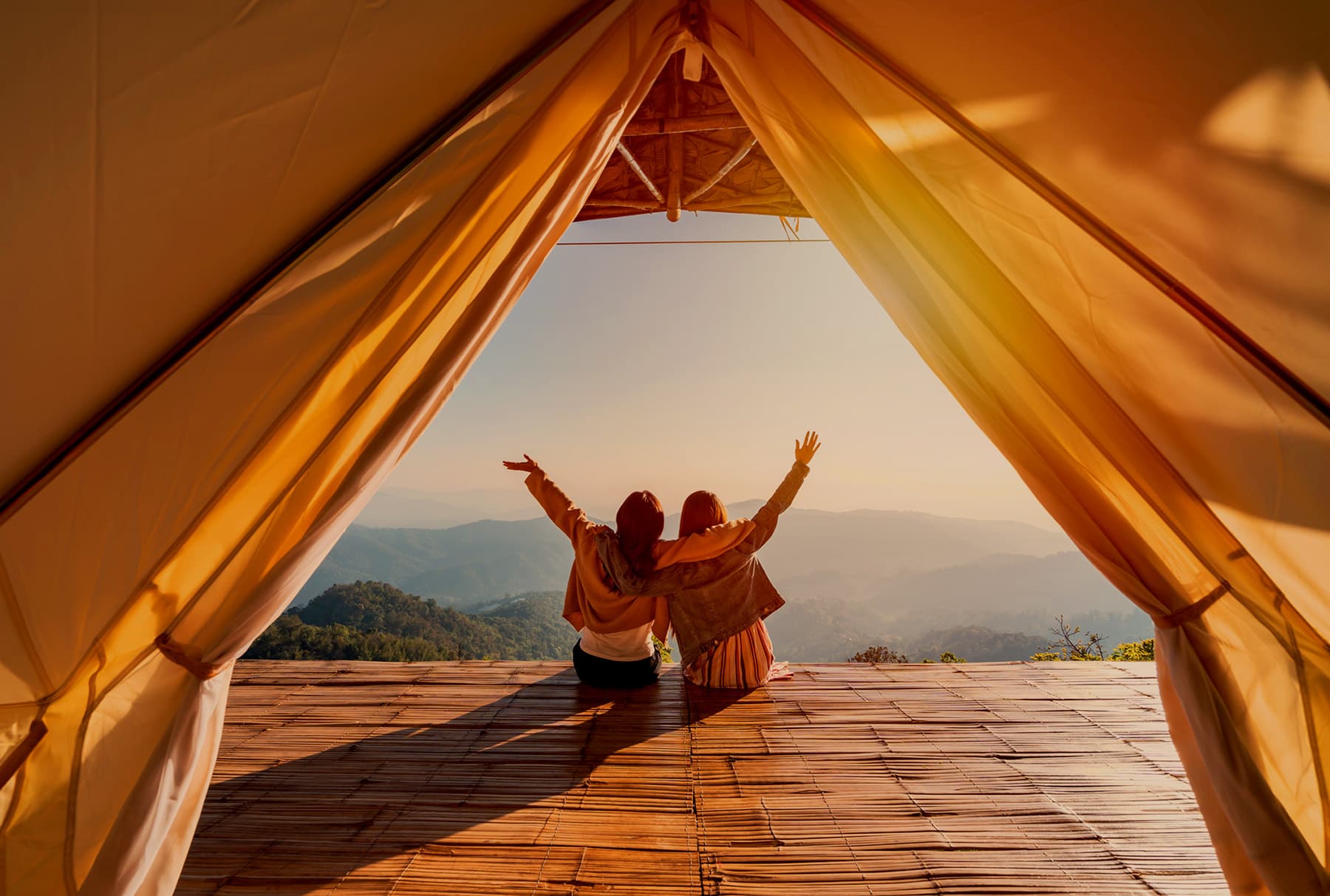 Two friends sitting arm in arm on a bamboo platform in the open mouth of a canvas tent, arms raised towards hazy blue mountains, the whole frame lit amber through the fabric